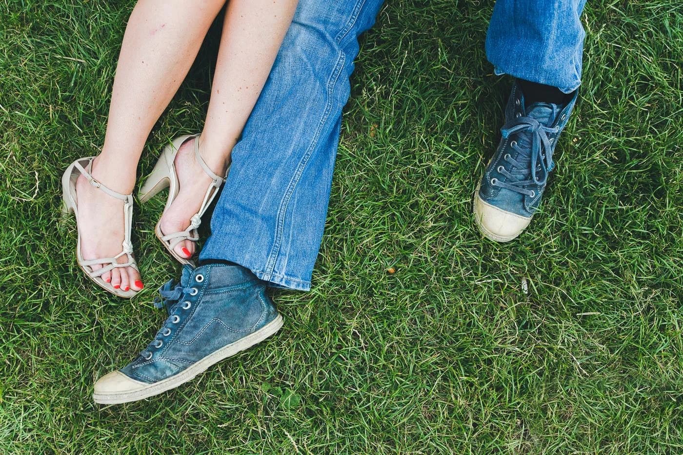 Twee paar voeten naast elkaar in het gras, van bovenaf gezien. Blote benen met beige sandalen met hak naast spijkerbroekpijpen met blauwe sneakers.