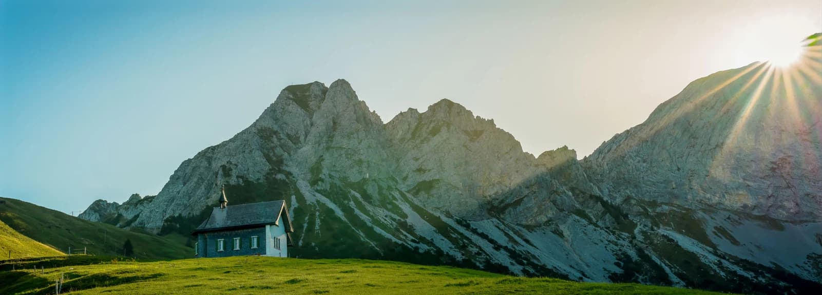 Een kapel op een alpenweide, met de bergkam en de opkomende zon erachter